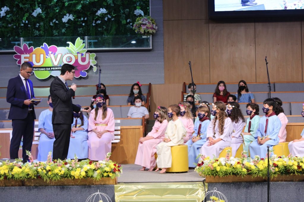 11 crianças usando becas batismais coloridas, sentadas e ouvindo a programação.