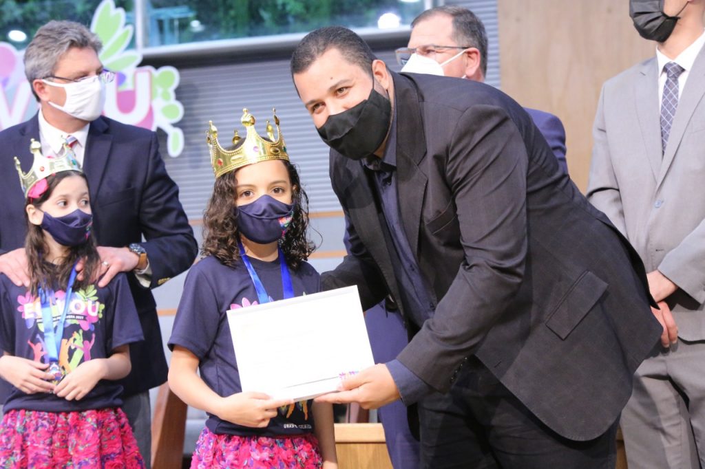 Menina de 8 anos recebendo certificado das mãos do diretor da escola.